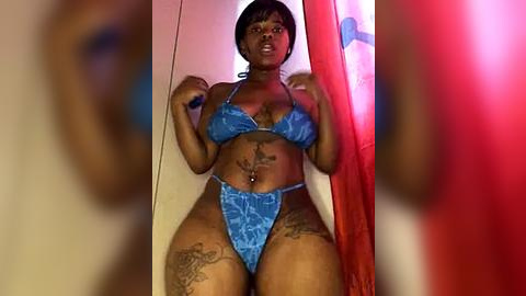 Media: Video of a curvy Black woman with short hair, wearing a blue floral bikini, standing in a doorway, with blurred figures in the background.