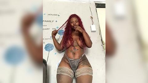 Media: Video of a nude, dark-skinned woman with long red hair, wearing lace thigh-high stockings and a silver thong, posing in a bathroom with white walls and blue dandelion decals.