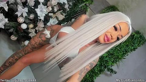 Media: Video of a fair-skinned woman with long, platinum blonde hair, wearing a white bikini, posing in front of a decorated Christmas tree. Her body is adorned with intricate tattoos.