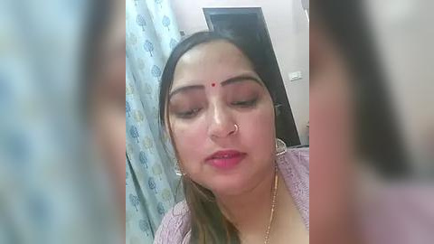 Media: A video of a young South Asian woman with fair skin, long dark hair, and red bindi, wearing a pink sweater, sitting in a bathroom with a floral-patterned shower curtain.