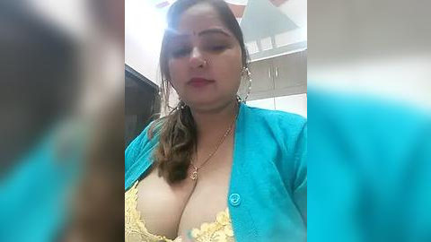 Media: A video of a woman with medium skin tone, wearing a turquoise jacket over a yellow lace bra, standing in a kitchen with white cabinets and a dark background.