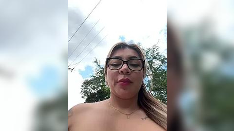 Media: A video of a topless Latina woman with medium skin tone and long brown hair, wearing glasses and a gold necklace, standing outdoors against a cloudy sky with trees in the background.