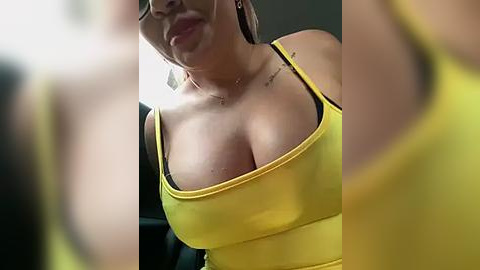 Media: Video of a curvaceous woman with light skin, large breasts, and a yellow tank top, viewed from below. Background is blurry, focusing on her chest and upper body.