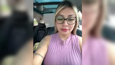 Media: A video of a light-skinned woman with blonde hair and glasses, wearing a sleeveless, light purple top, sitting in a car with a blurred background.