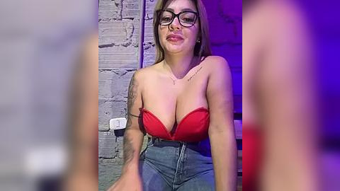 Media: Video of a woman with glasses, large breasts, and tattoos, wearing a red bra and high-waisted jeans, seated against a brick wall in a dimly lit room.