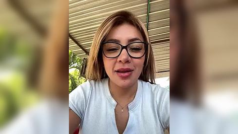 Media: Video of a young, light-skinned woman with straight, shoulder-length brown hair and large, black-framed glasses, wearing a white, short-sleeved shirt, sitting outdoors under a beige awning.