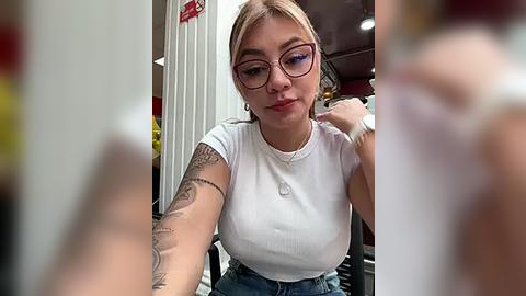 Media: Video of a young woman with light skin, blonde hair, and glasses, wearing a white ribbed tank top and blue jeans, sitting indoors. She has a tattoo on her left arm and a watch on her wrist.