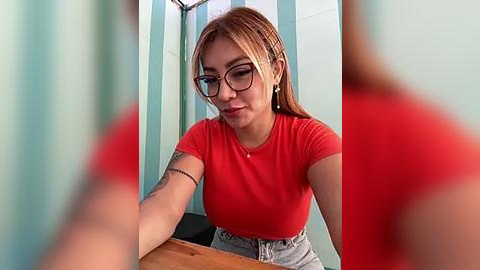 Media: A video of a Latina woman with light skin, straight, shoulder-length brown hair, wearing glasses, a red T-shirt, and high-waisted jeans, seated at a wooden table in a minimalist room.