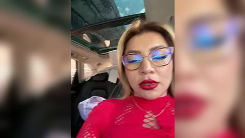 Media: A video captures a woman with fair skin, long blonde hair, and blue eyeshadow, wearing red lipstick and a red, lace-top dress. She sits in a car with tinted windows, visible through the driver's side window.