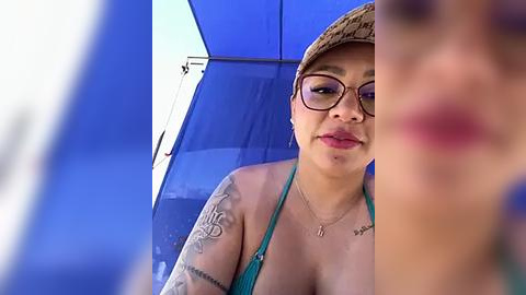 Media: Video of a light-skinned woman with glasses, wearing a turquoise bikini and a woven hat, under a blue umbrella, tattoos visible.