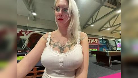 Media: Video of a blonde woman with glasses and red lipstick, wearing a white tank top, standing in a dimly lit room with pool tables and colorful graffiti on the walls.