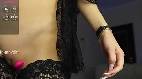 Media: Video of a light-skinned woman with a flat stomach, wearing black lace lingerie and a wristwatch, holding a pink vibrator. Background shows a dimly lit room.