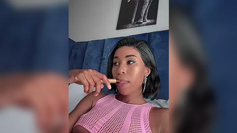 Media: Video of a Black woman with short, dark hair and medium skin tone, applying lipstick with a golden brush, wearing a pink mesh top. Background features a blue wall and a black-and-white framed video.