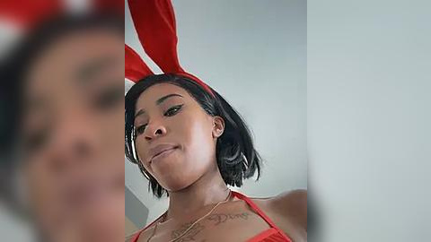 Media: A video of a young woman with a short black bob haircut wearing a red bunny headband and a red halter top, with a blurred figure in the background.