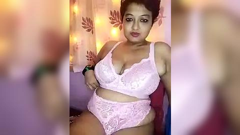 Media: Video of a curvy, dark-skinned woman with medium-length hair, wearing light pink lace lingerie. She's seated in a cozy room with warm, yellow string lights and floral curtains. Her expression is confident.