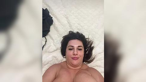 Media: Video of a fair-skinned woman with dark hair lying on a white, textured blanket, wearing a black bra. Her breasts are partially visible.