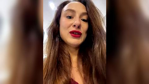 Media: Video of a woman with long brown hair and red lipstick, wearing a red top, taken from a close-up angle, with blurred background.