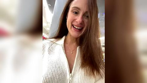 Media: Video of a smiling, fair-skinned woman with long, straight brown hair, wearing a white ribbed knit top with a zipper, and a delicate necklace.