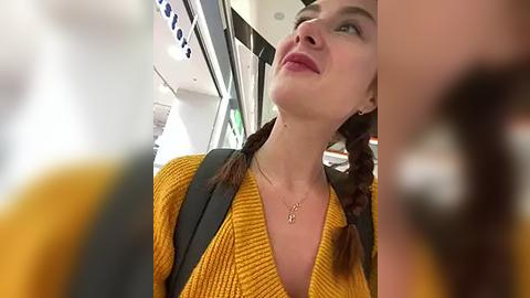 Media: Video of a young woman with fair skin, brown hair braided, wearing a mustard-yellow knit top, gazing upward, seated on a train. Background shows train interior with black seats and overhead compartments.