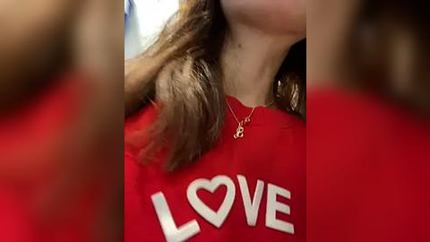 Media: Video of a woman wearing a red shirt with \"LOVE\" in white letters. Her long, wavy brown hair is partly visible. She wears a delicate gold necklace with a small heart pendant. Background is blurry.