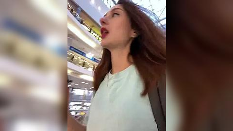 Media: Video of a woman with long, straight brown hair, wearing a light blue blouse, shopping in a brightly lit supermarket. Shelves with various products are visible in the background.