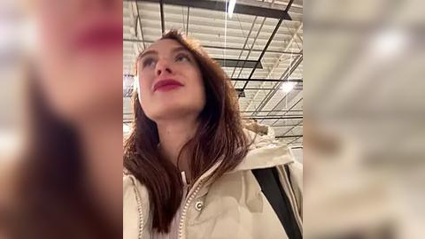 Media: Video of a young woman with fair skin, long brown hair, and red lipstick, wearing a cream-colored jacket, standing in a bright, modern store with a high ceiling and industrial lighting.