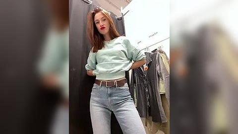 Media: Video of a young woman with long brown hair, wearing a light green sweater and high-waisted light blue jeans, standing in a brightly lit clothing store with hanging garments in the background.