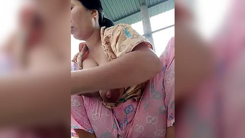 Media: A video of an Asian woman with medium skin tone and black hair, wearing a pink floral-patterned shirt, exposing her breast, crouching indoors with a corrugated metal ceiling.