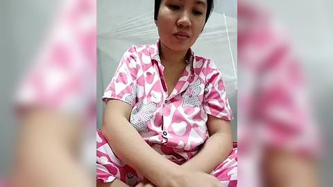 Media: Video of a woman in pink and white patterned pajamas, sitting with her hands clasped, in a white room with a tiled ceiling.