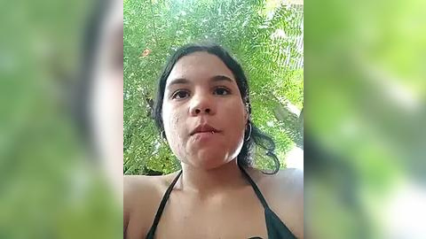 Media: Video of a young woman with medium skin tone, dark hair, and a neutral expression, wearing a black bikini top. She is surrounded by lush green foliage, suggesting a garden or outdoor setting.