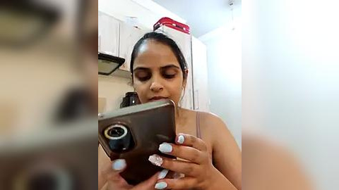 Media: A candid video of a woman with medium skin tone and straight dark hair in a ponytail, taking a selfie in a small, cluttered kitchen. She's wearing a tank top and has light blue nail polish. The background features white cabinets, a red container, and a stove.