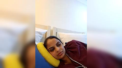 Media: Video of a woman with dark hair, wearing a maroon jacket, lying on a bed with white pillows and a blue blanket. She has a yellow pillow over her head, appearing to sleep or rest. The room is brightly lit.