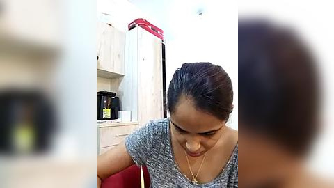 Media: Video of a young woman with medium brown skin and dark hair in a ponytail, wearing a gray sweater, leaning forward in a modern kitchen with light-colored cabinets, a red oven, and a black coffee machine.