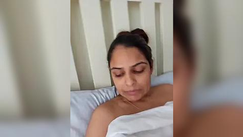 Media: Video of a young woman with medium brown skin, brown hair tied back, lying on a bed with white sheets, wearing a light-colored top, eyes closed, looking relaxed.