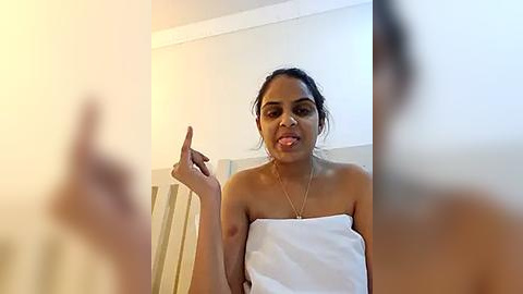 Media: Video of a young woman with medium-brown skin and dark hair, wearing a white towel, sticking out her tongue and making a finger gesture.