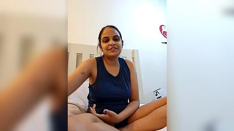 Media: A video of a smiling South Asian woman in a dark blue sleeveless top, sitting on a bed in a simple, brightly lit bedroom.