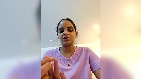 Media: Video of a young South Asian woman with light skin and dark hair, wearing a lavender scrub top, holding a small object in her hand. The background is a minimalist, off-white room with a soft light.