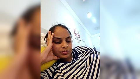 Media: A video featuring a young woman with medium brown skin, wearing a blue and white striped shirt, sitting in a brightly lit room with white walls and a ceiling fan.