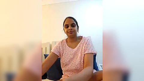 Media: A video of a South Asian woman with medium skin tone, wearing a light pink floral dress, sitting on a bed in a dimly lit room.