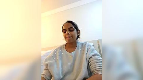 Media: Video of a South Asian woman with medium skin tone, wearing a light gray sweater, with short dark hair, sitting on a white bed, eyes closed, serene expression.