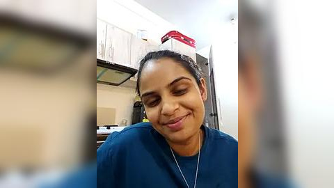 Media: Video of a smiling, dark-skinned woman in blue scrubs, wearing a white cap, standing in a kitchen with white cabinets and a red box on top.