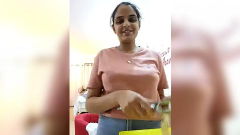 Media: Video of a smiling South Asian woman with medium brown skin, dark hair in a ponytail, wearing a pink t-shirt and blue jeans, holding a yellow envelope in a dimly lit room.