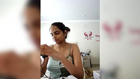 Media: Video of a young South Asian woman with medium skin tone, dark hair in a bun, wearing a light gray spaghetti-strap tank top, sitting on a bed in a dimly lit bedroom.