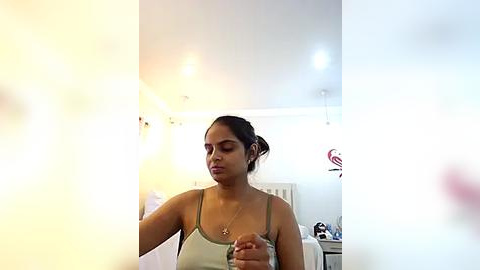 Media: Video of a young woman with medium brown skin and dark hair in a bun, wearing a green tank top, standing in a brightly lit bedroom with white walls, a bed, and a window.