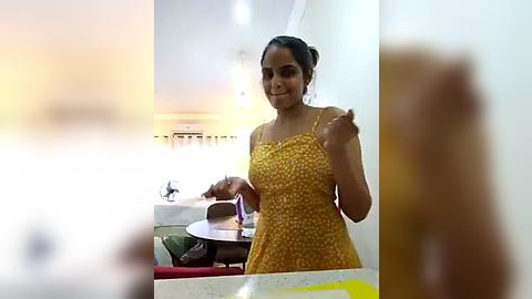 Media: Video of a young South Asian woman with medium skin tone and dark hair in a bun, wearing a yellow floral dress, gesturing in a modern, brightly lit room with blurred background.