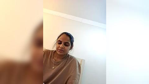 Media: Video of a woman with medium skin tone and dark hair in a side ponytail, wearing a beige sweater, sitting indoors against a white wall.