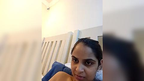 Media: A blurry video of a young South Asian woman with dark hair, lying on a bed with white and blue bedding, smiling into the camera.