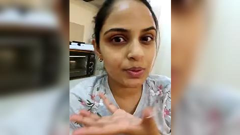 Media: Video of a young South Asian woman with dark hair, medium skin tone, and brown eyes, wearing a light floral top, smiling, and gesturing with both hands in a kitchen with a microwave and stove in the background.