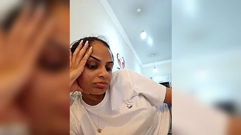 Media: Video of a young woman with medium brown skin, wearing a white t-shirt, sitting on a couch, touching her forehead in a concerned expression. Background shows a bright, modern room with white walls and recessed lighting.