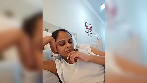 Media: Video of a young woman with medium-brown skin, resting on a bed, wearing a white shirt, with a contemplative expression.